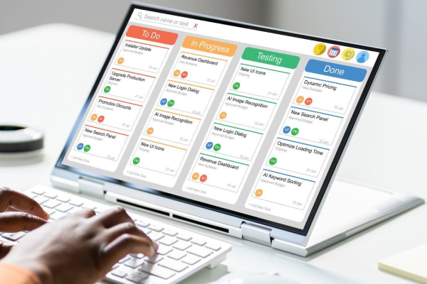 Hands using a white keyboard with a medium sized monitor that has a project management software on it, and a white mouse all on a white table for project management software (project management tool)